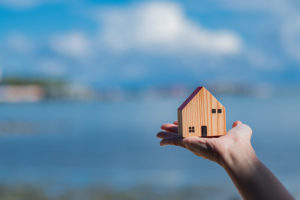Wooden house held in hand in front of blue sky and water background