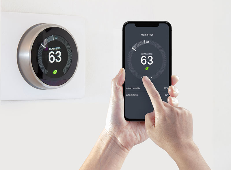 Hand holding a smart phone next to a wall mounted thermostat, setting the temperature of the thermostat via the smart phone.