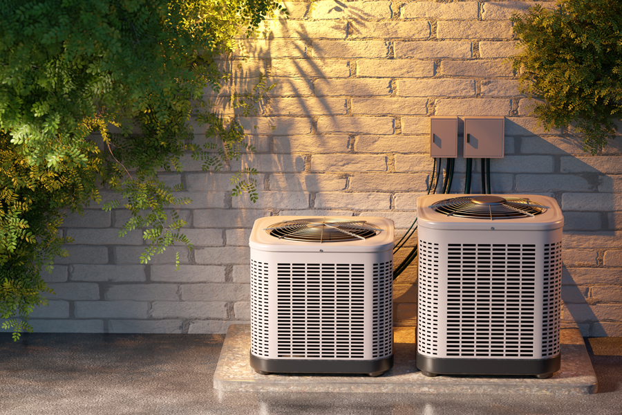 Two outdoor air conditioning units installed on a concrete base against a brick wall with greenery around
