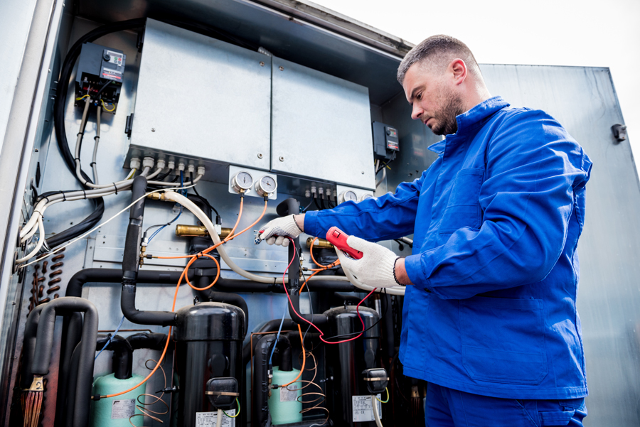 A technician in a blue uniform and gloves uses a multimeter to check electrical connections inside an open industrial control panel.