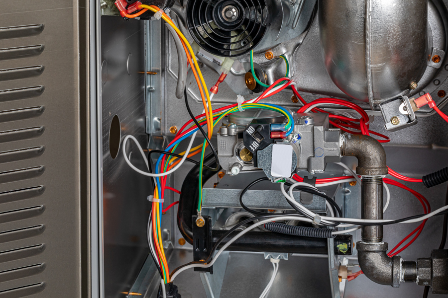 Close-up view of the internal components and wiring of a residential gas furnace, showing pipes, wires, a fan, and various mechanical parts.