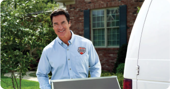 A service worker in a blue shirt stands outside a house, holding a large gray item near a white van. Green trees and a brick house are visible.
